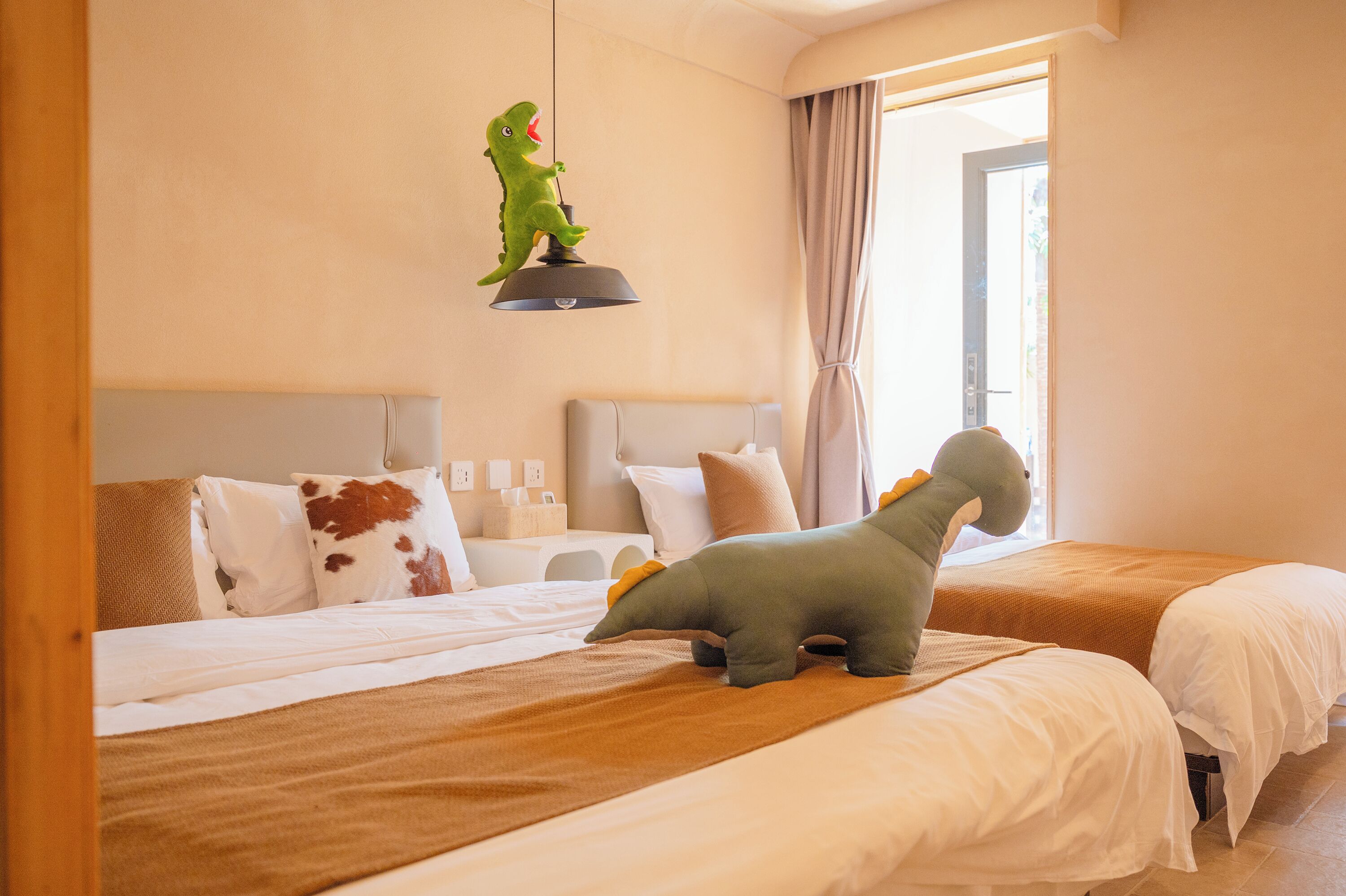 A hotel room with two beds, a dinosaur toy on the left bed, a hanging lamp, and a window with curtains.