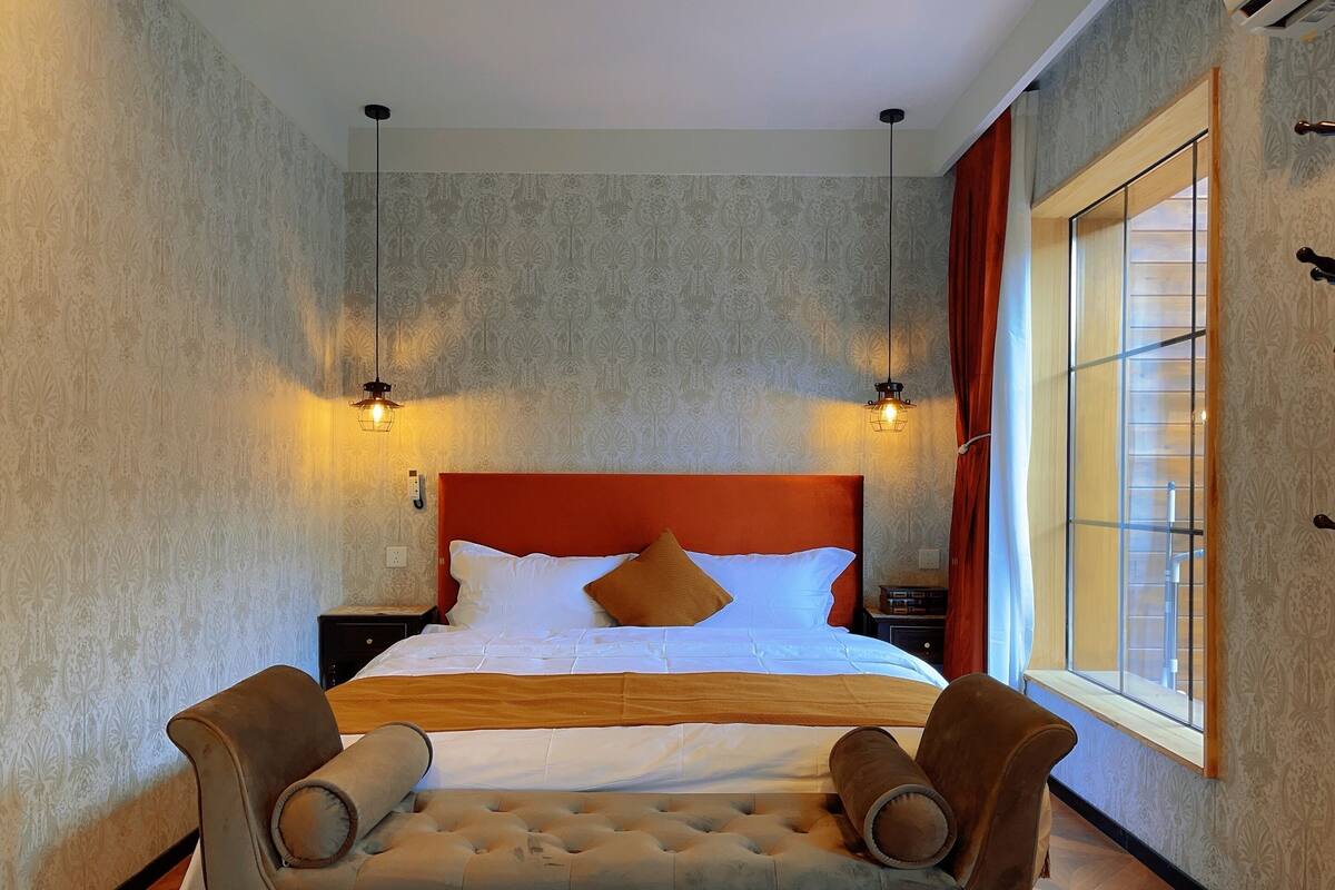 A hotel room with a large bed, two bedside lamps, a window with curtains, and a tufted sofa.