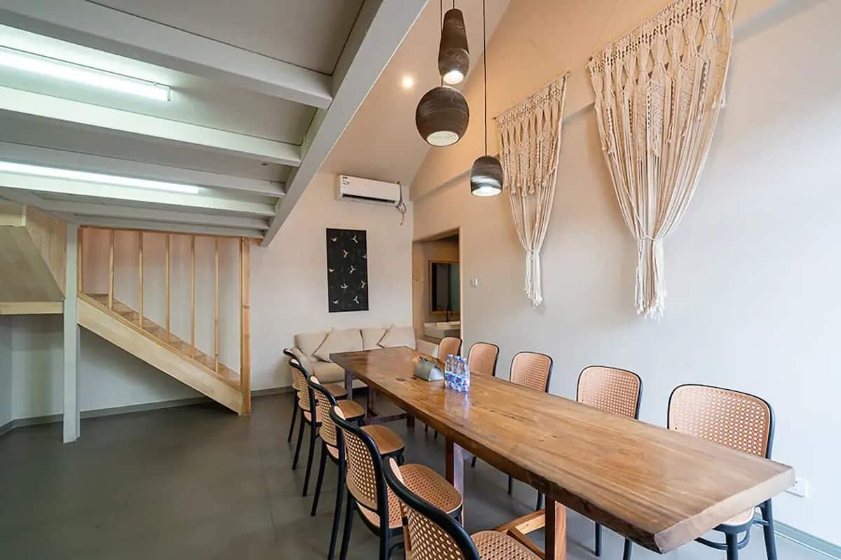 A dining area with a long wooden table, wicker chairs, pendant lighting, macrame wall hangings, and a staircase.
