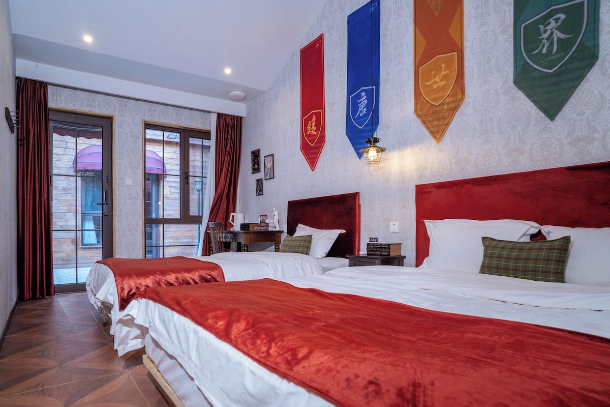 A hotel room with two beds, a desk with a chair, a window with curtains, and flags on the wall.