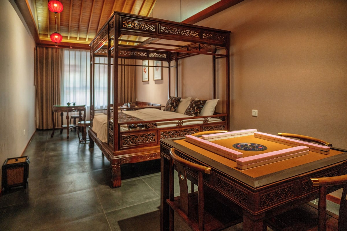 A traditional four-poster bed with intricate woodwork, a tatami mat game table, and a small wooden table with a chair in a room with a sloped ceiling and wooden beams.