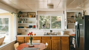 A kitchen with wooden cabinets, a dining table with chairs, a refrigerator, and a window with a view of greenery.