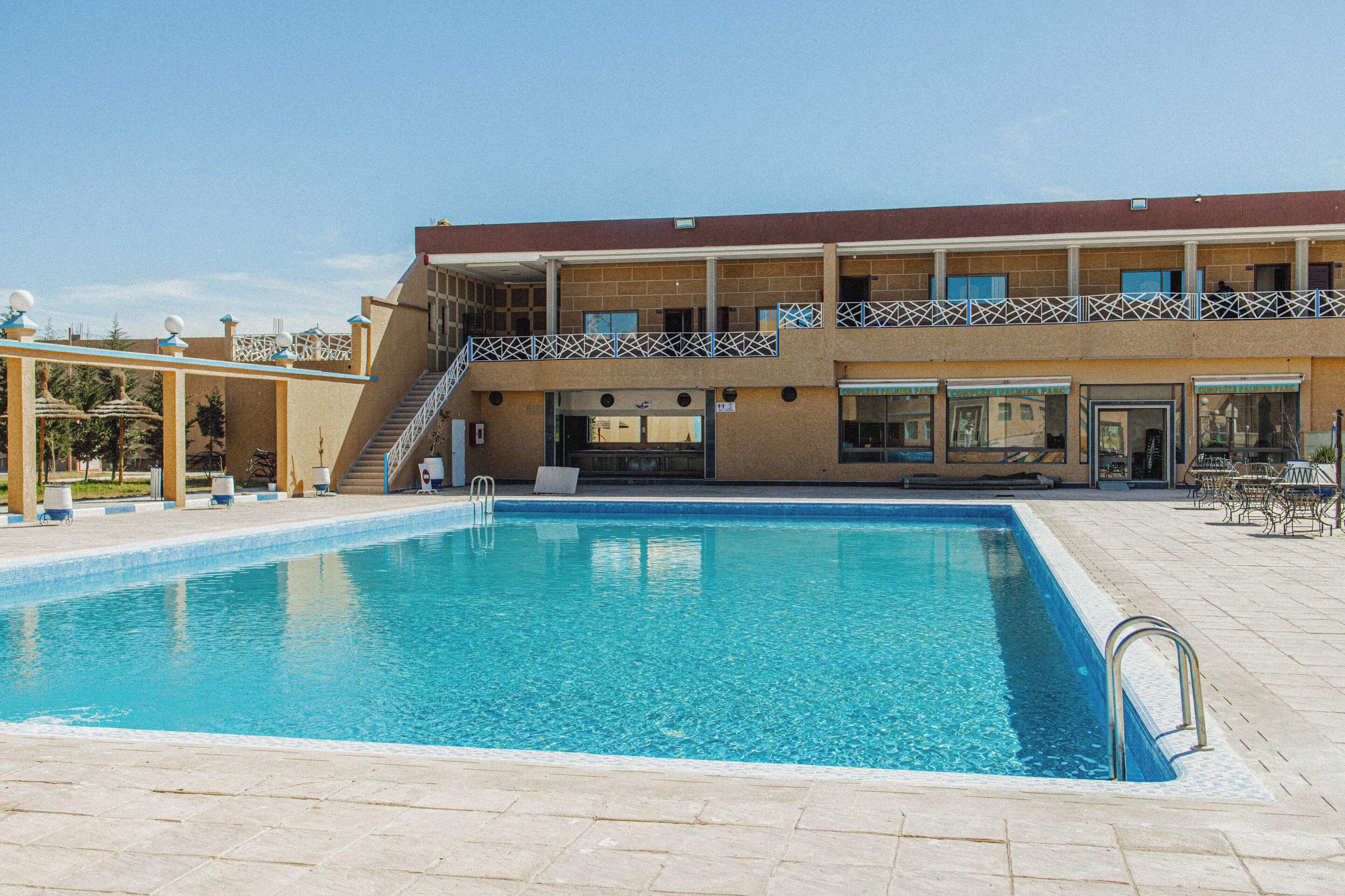 Una piscina rectangular con una escalera de metal, una zona cubierta con techo y un edificio de dos pisos con balcón.