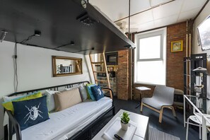 Een knus woongedeelte met een bed, een klein tafeltje, een stoel, een kamerplant en een boekenplank.