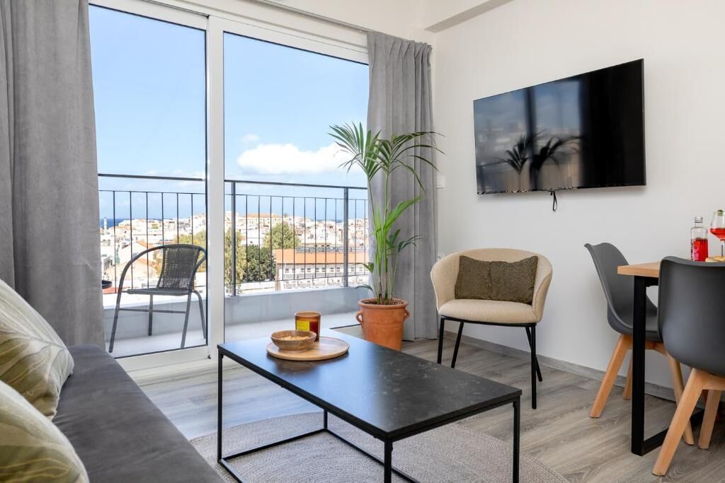 A modern living room with a sofa, a dining table with chairs, a potted plant, a flat-screen TV, and a balcony with a view of the city.