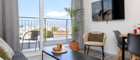 A modern living room with a sofa, a dining table with chairs, a potted plant, a flat-screen TV, and a balcony with a view of the city.