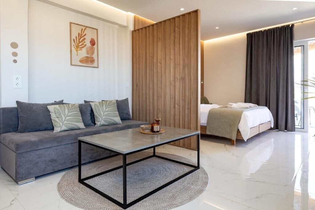 A modern hotel room with a grey sofa, a small round coffee table, a bed with a white duvet, and a wooden partition.