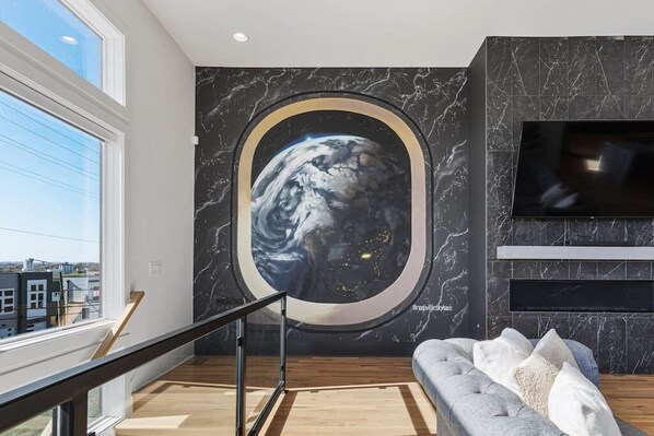 A modern living room with a large window, a wall-mounted TV, and a circular artwork of Earth.