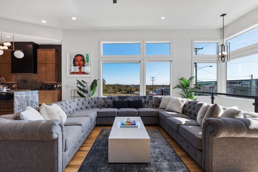 A modern living room with a grey sectional sofa, a coffee table, and large windows offering a view of the outdoors.