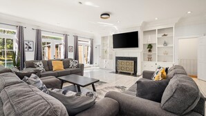A spacious living room with a large sofa set, a flat-screen TV mounted above a fireplace, and a view of the outdoors through large windows. - AL Oasis | Sleeps 12 | Hot Tub | Sauna | Pool Table (Mobile)
