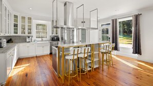 A modern kitchen with white cabinetry, a central island with bar stools, stainless steel appliances, and a large window with curtains. - AL Oasis | Sleeps 12 | Hot Tub | Sauna | Pool Table (Mobile)