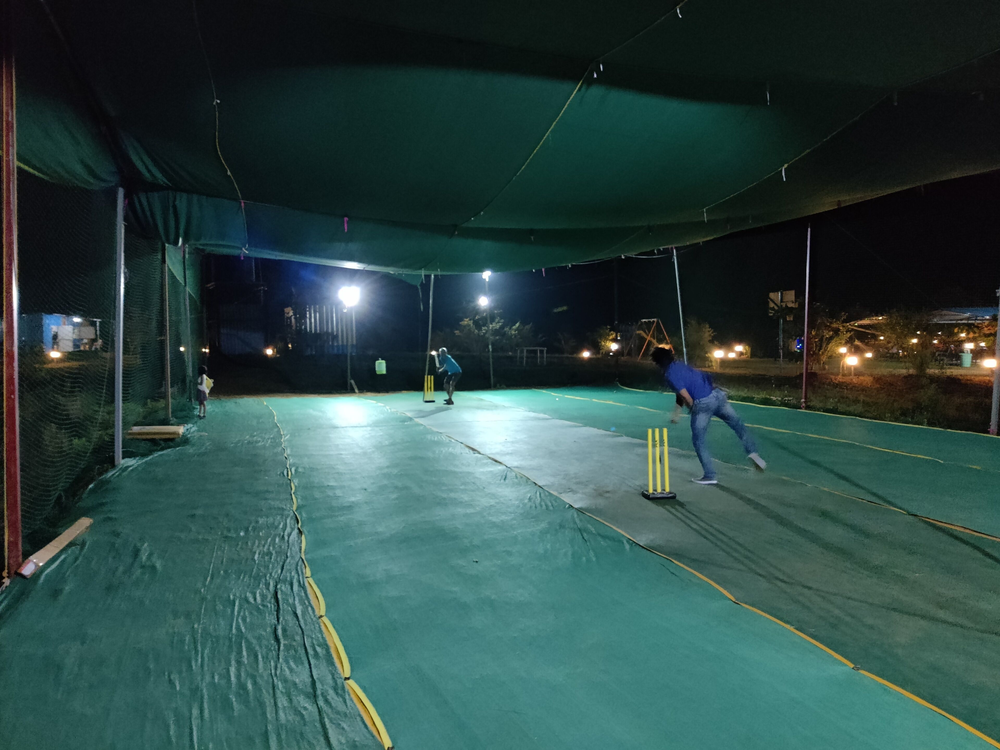 Een cricketveld buiten met nachtverlichting, een omheining van netten en een groen speeloppervlak.