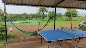 Un espace extérieur abrité comprenant une table de ping-pong, un hamac et une vue sur un champ verdoyant, avec des arbres et un plan d’eau en arrière-plan.