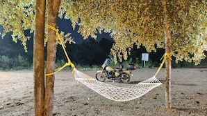Un hamac tendu entre deux arbres, une moto garée à proximité et un sol en terre battue.