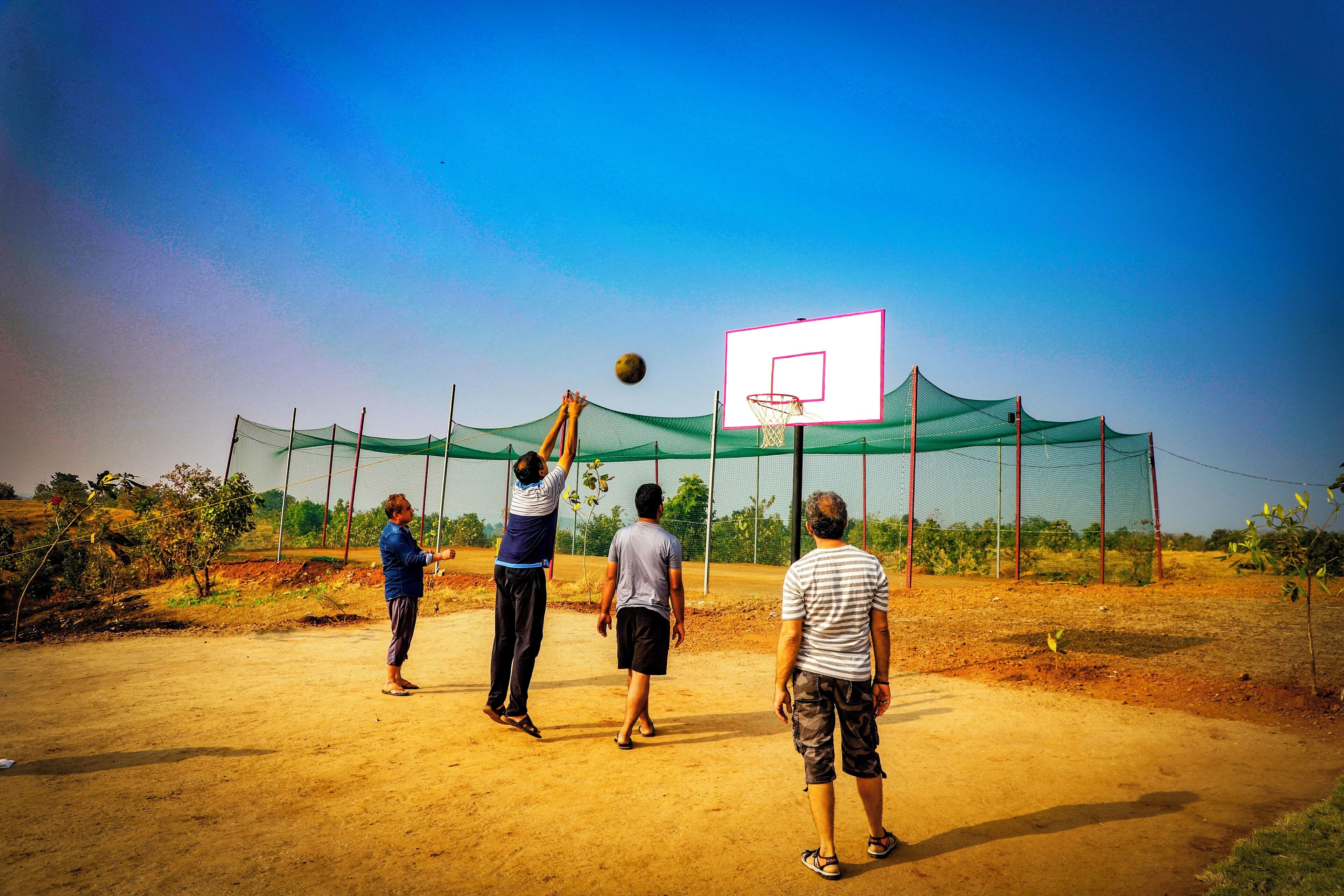 A group of people playing basketball on a dirt court with a netted enclosure and a basketball hoop.