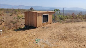 Une petite cabane en bois, surmontée d’une antenne parabolique, entourée d’herbes sèches et de quelques arbres clairsemés, avec des montagnes en arrière-plan.