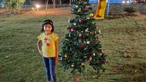 Un enfant se tient à côté d’un sapin de Noël décoré, dans un espace extérieur enherbé, la nuit.