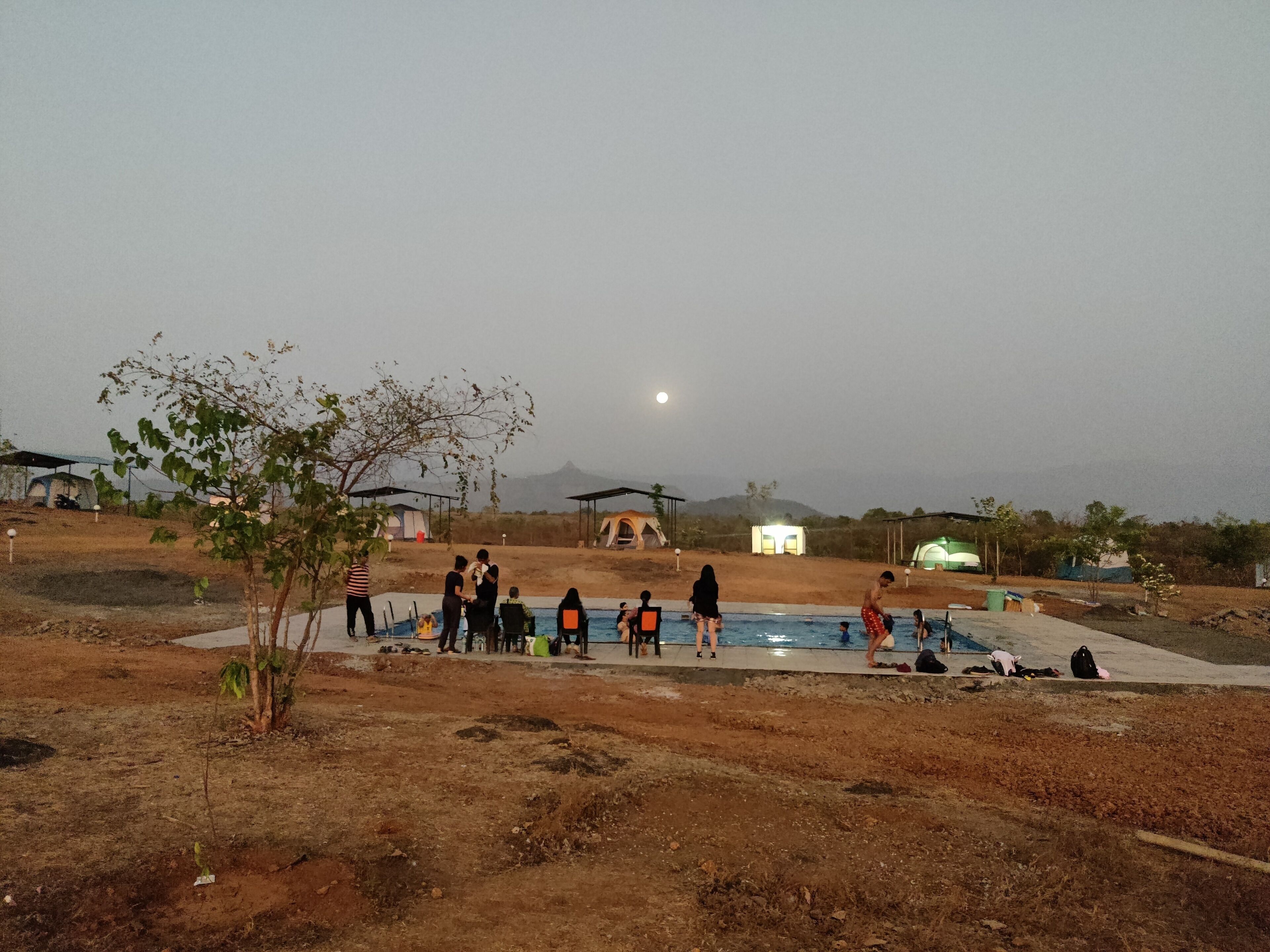 A group of people around a swimming pool in a rural setting with tents and a mountain in the background.