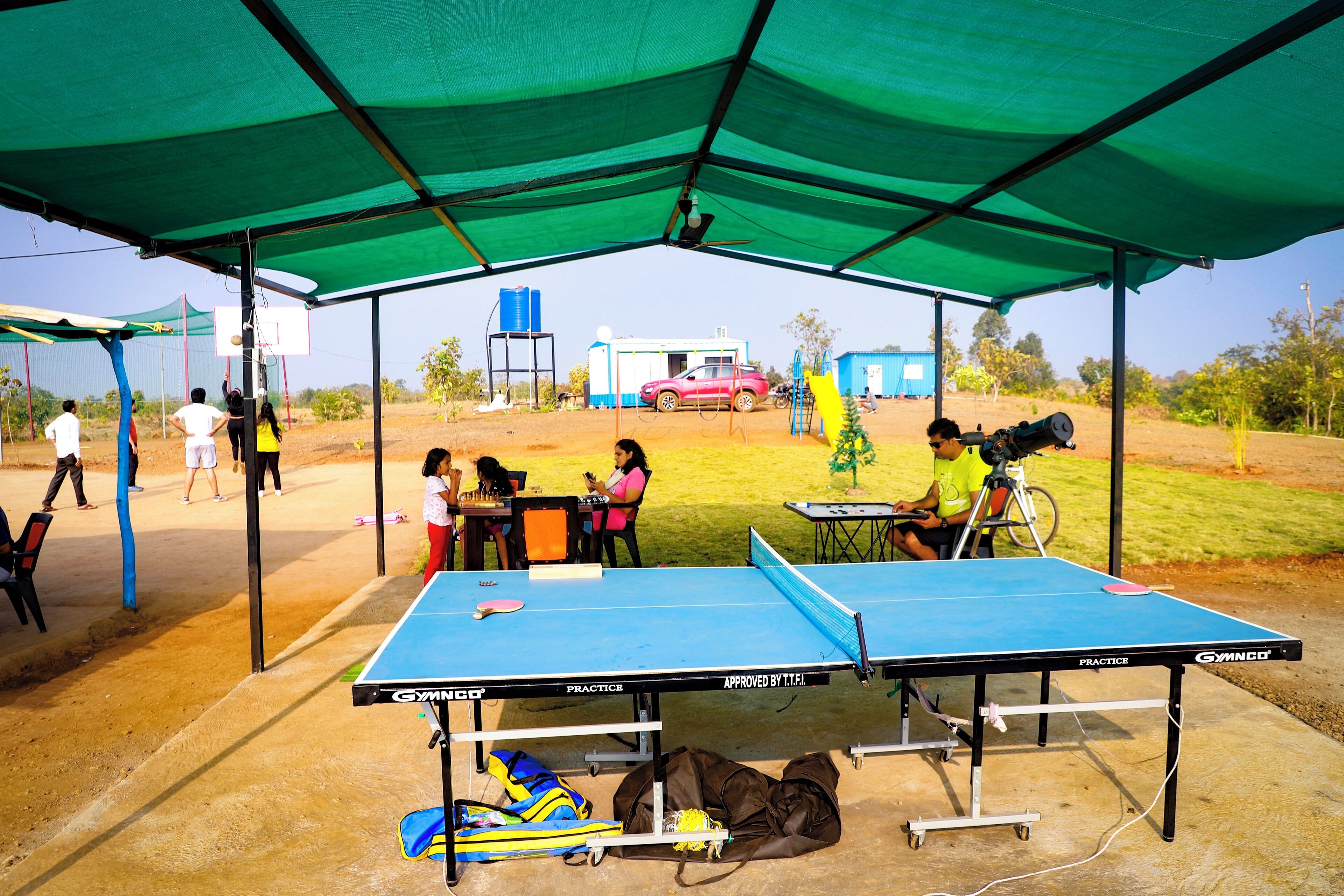 An outdoor table tennis setup with a canopy, a blue table, and a red car in the background.