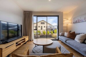 Ein modernes Wohnzimmer mit Flachbildfernseher, rundem Tisch, einer Couch und Blick auf ein Gebäude sowie einen geparkten Wagen durch eine Glastür.