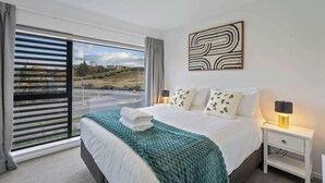 Een moderne hotelkamer met een groot bed, uitzicht op een weg en heuvels, een schilderij aan de muur en een nachtkastje met een lamp.