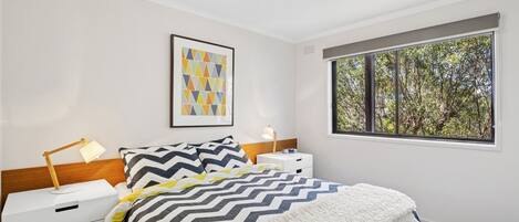 A bedroom with a bed, a bedside table with a lamp, a nightstand, a framed picture on the wall, and a window with a view of trees.