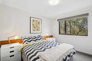 Ein Schlafzimmer mit Bett, Nachttisch mit Lampe, ein Fenster mit Blick auf Bäume und einem Bild an der Wand.