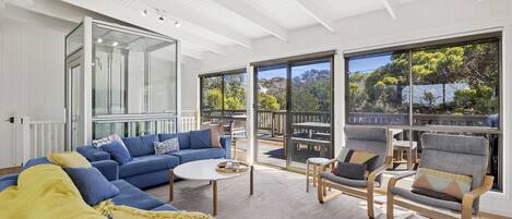 Ein modernes Wohnzimmer mit einem blauen Sofa, einem runden Couchtisch und grauen Sesseln. Eine Glastür führt zu einem Balkon mit Blick auf Bäume.