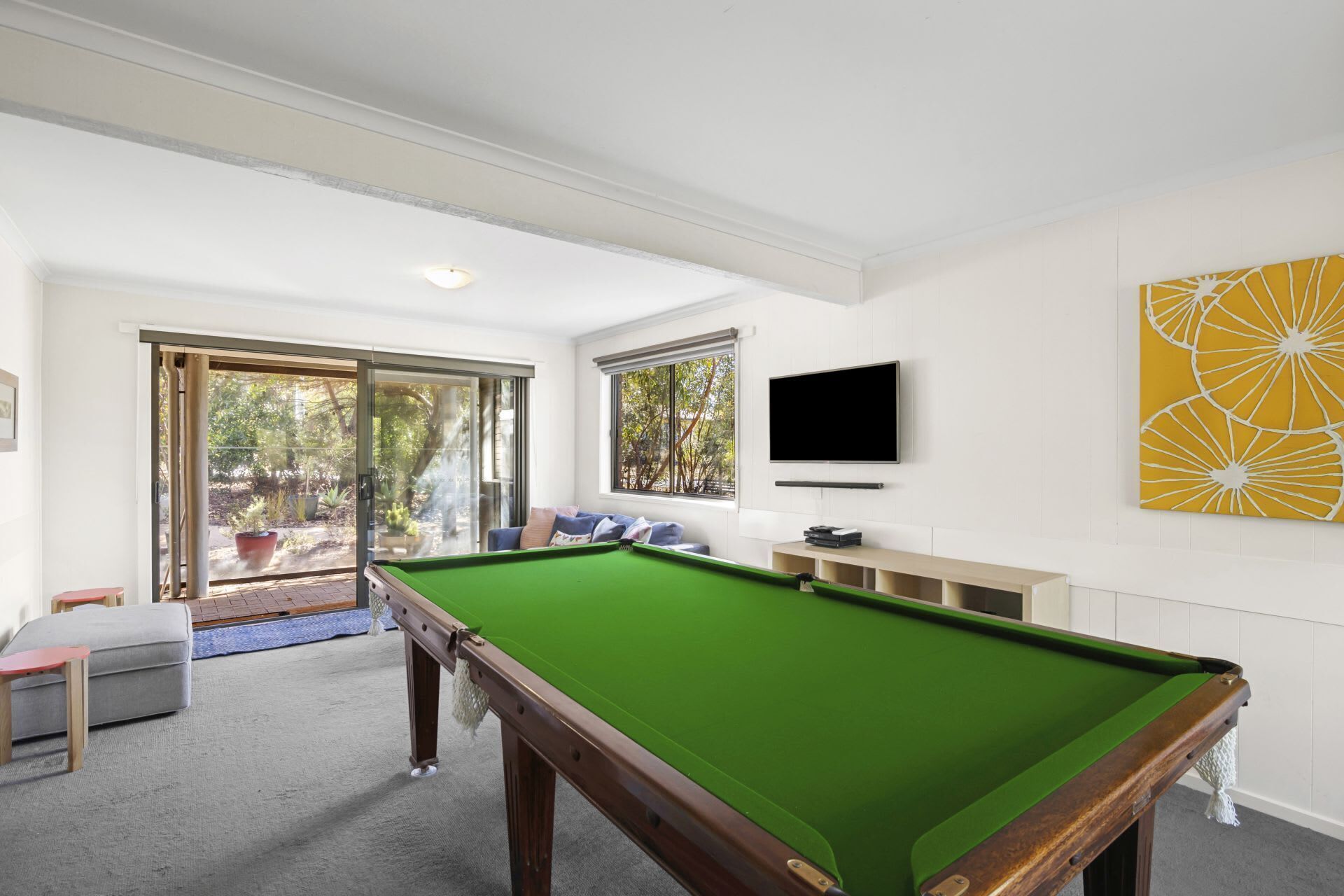 A pool table in a room with a sliding glass door leading to a garden, a flat-screen TV mounted on the wall, and a yellow abstract painting.
