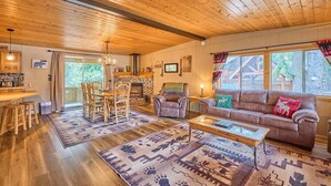 A cozy cabin interior with a wooden ceiling, a dining area with chairs, a sofa, and a fireplace.