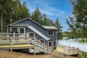 Une petite cabane dotée d’une terrasse et d’un escalier menant à une porte, située au bord d’un lac entouré d’arbres.