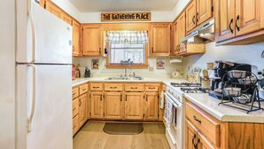Une cuisine avec des meubles de rangement en bois, un réfrigérateur, une cuisinière et une fenêtre agrémentée d’une lambrequin à carreaux.