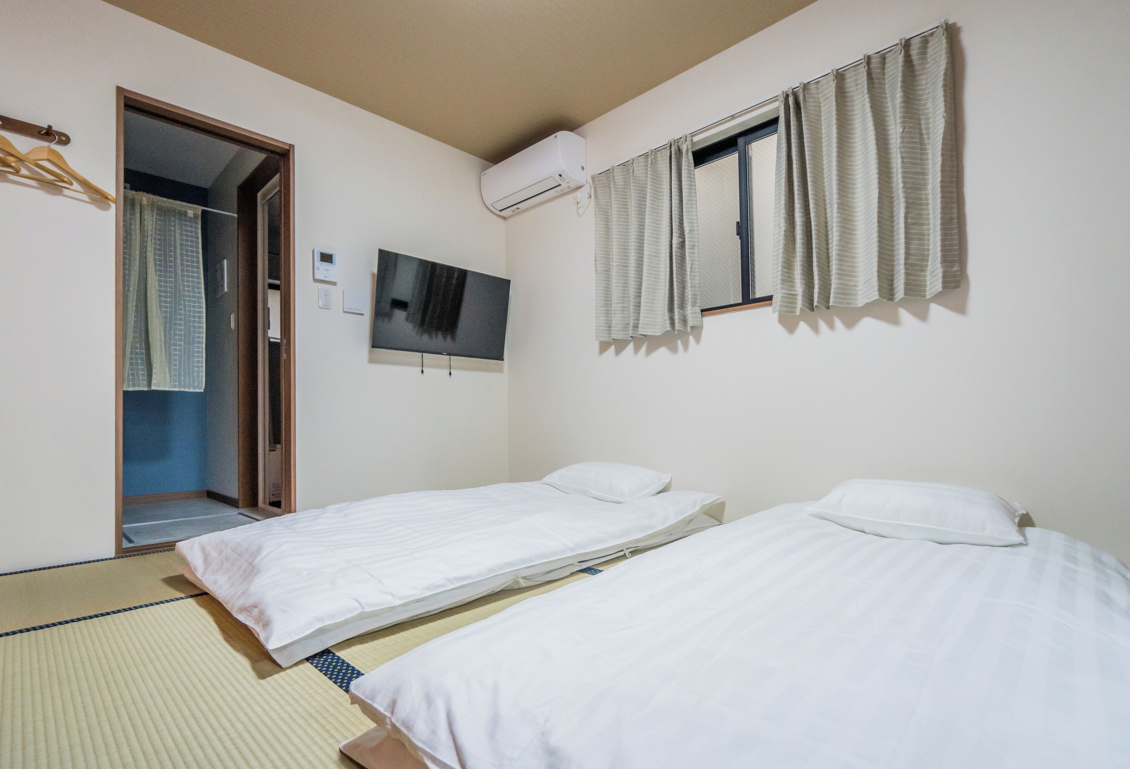 A hotel room with two single beds, a television mounted on the wall, an air conditioning unit, and a window with curtains.