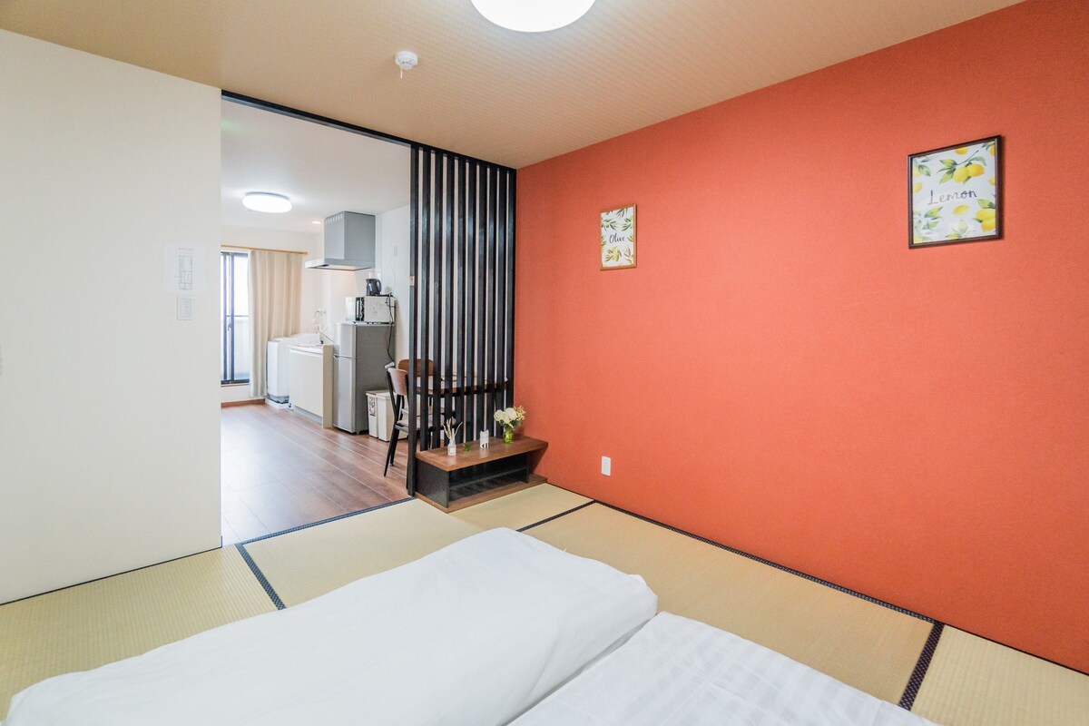 A hotel room with a bed, a wooden table with a vase, and a red wall with a lemon picture.