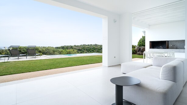 A modern living room with a white sofa, a small round table, and a TV. There is a large window offering a view of a swimming pool and greenery. - Villa 'Domo La Vela' with Sea View, Wi-Fi and Air Conditioning (Orosei)