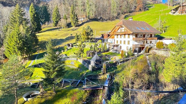 Ein großes Haus mit schrägem Dach, umgeben von üppiger, grüner Landschaft und einem kleinen Teich.