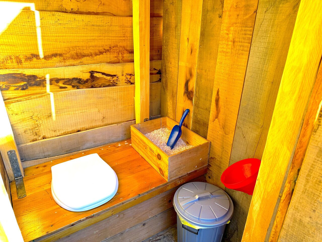 A wooden cabin with a wooden toilet, a blue shovel, and a red bucket.