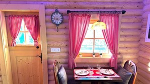 Ein Holzblockhaus-Interieur mit einem für zwei Personen gedeckten Esstisch, zwei Stühlen, einer Wanduhr und einem Fenster mit roten und weißen Karottenvorhängen.