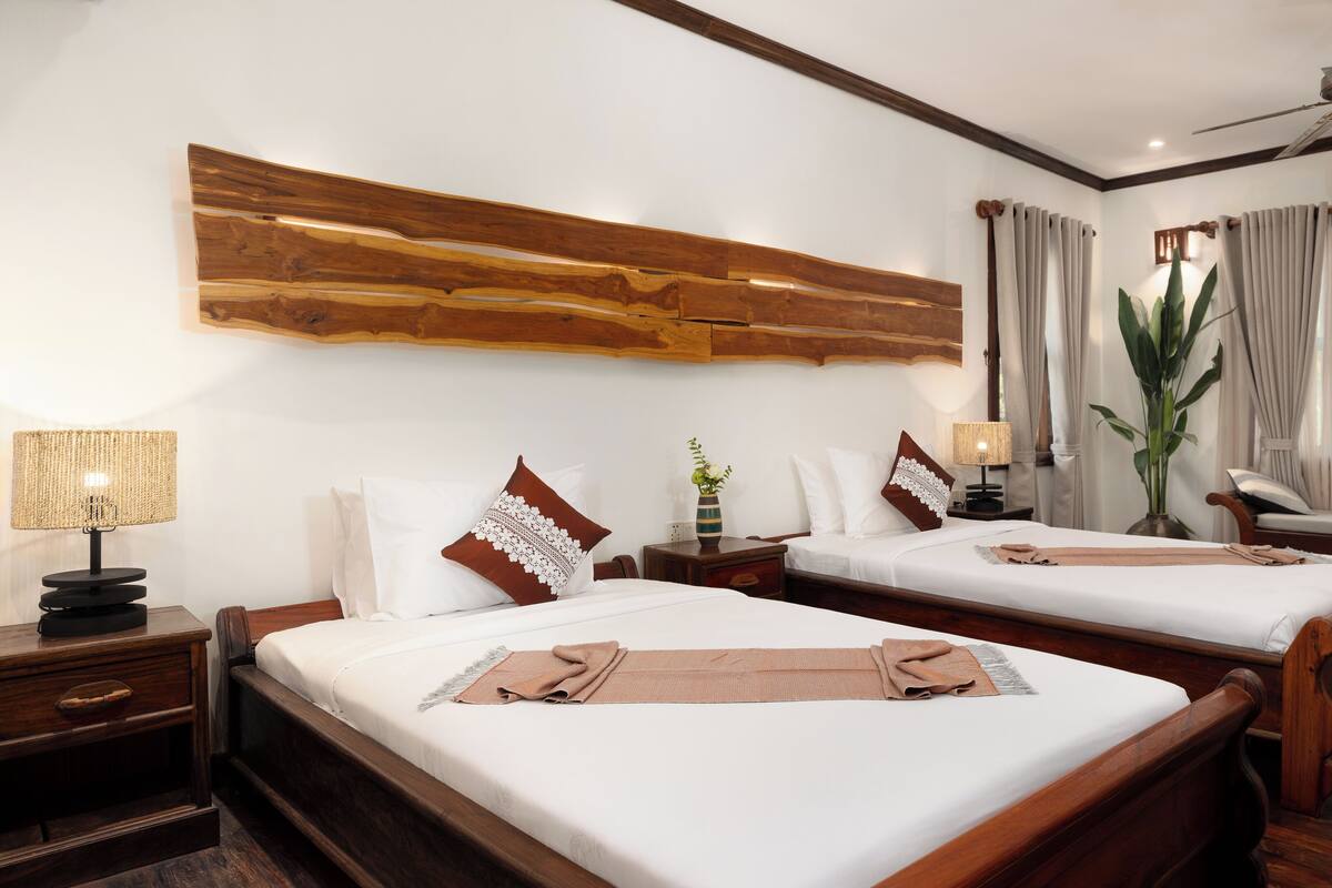 A hotel room with two beds, wooden headboards, a large wooden wall art piece, a ceiling fan, an air conditioning unit, a window with curtains, and a potted plant.