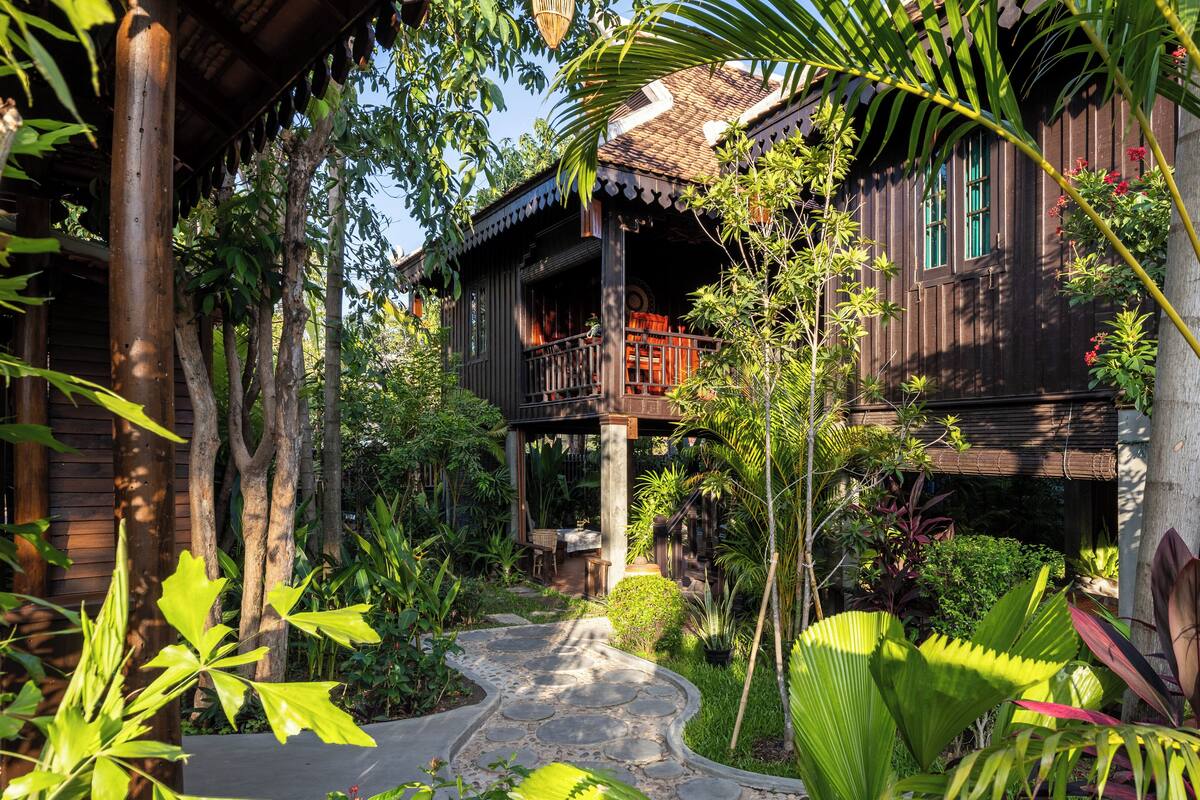 A traditional wooden house with a balcony, surrounded by lush greenery and a stone pathway.
