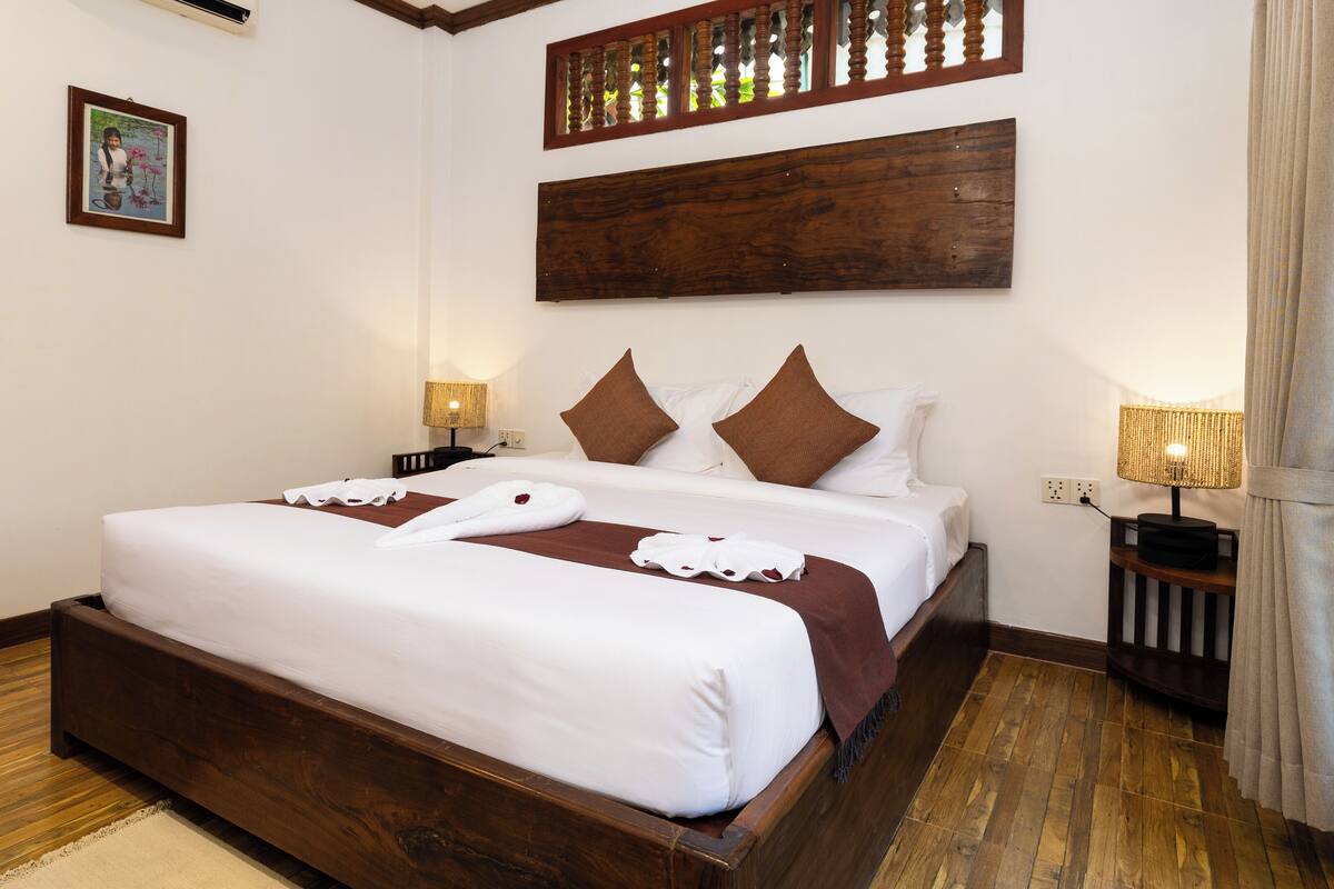A neatly made bed with white linens and brown accents, a wooden headboard, and a nightstand with a lamp.