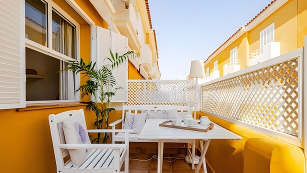 Ein Balkon mit einem weißen Tisch, einem Stuhl, einer Topfpflanze und einem weißen Gitter.