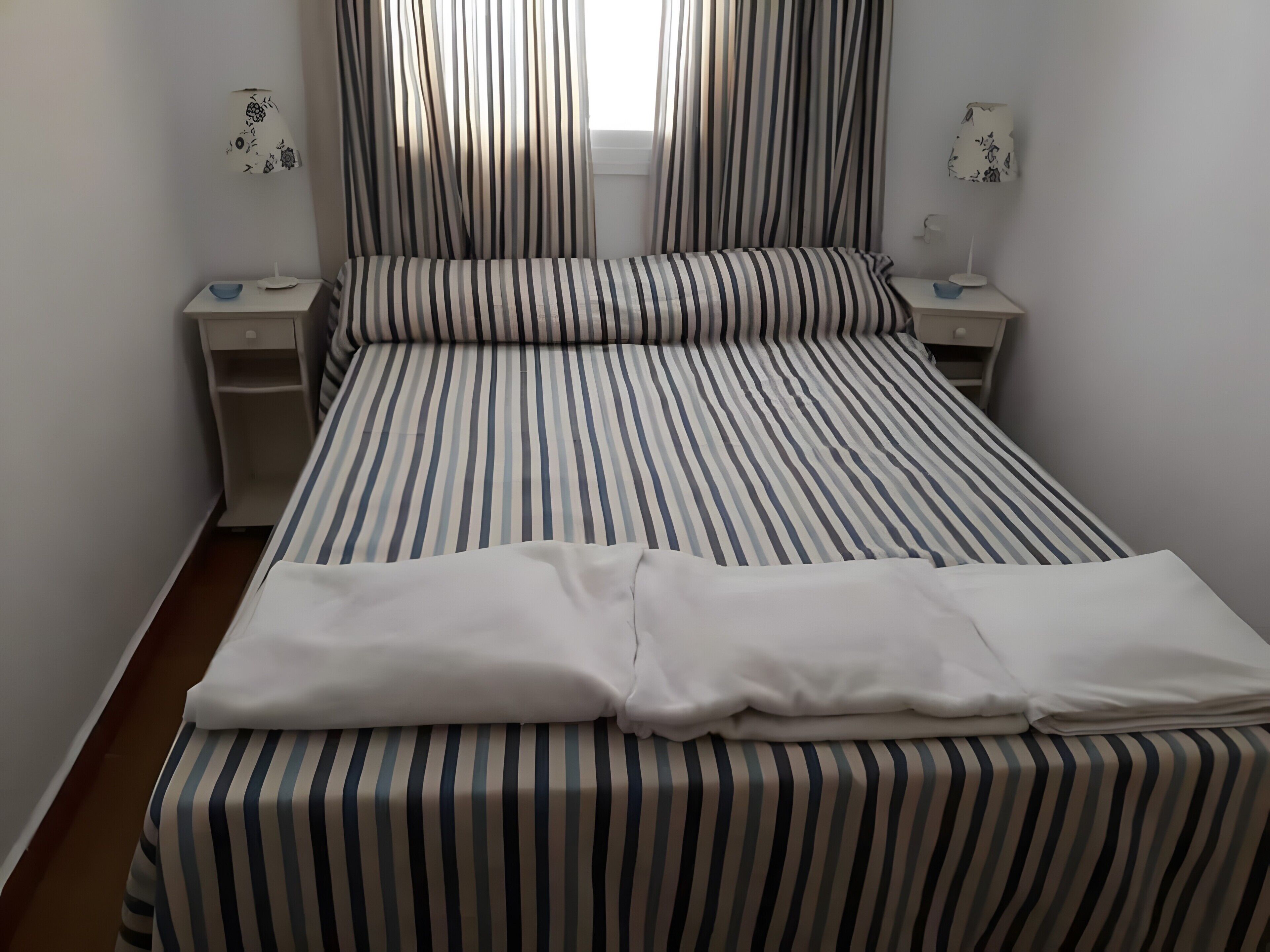 Una cama con ropa de cama a rayas, dos mesitas de noche con lámparas y una ventana con cortinas.