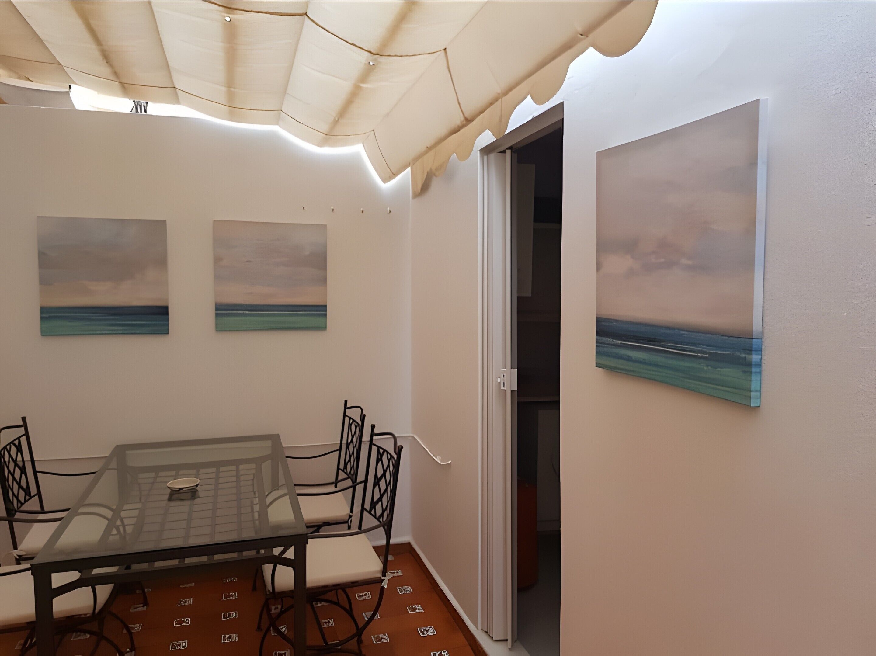 Un coin repas avec une table et des chaises, deux tableaux représentant la mer, et une porte donnant sur une autre pièce.