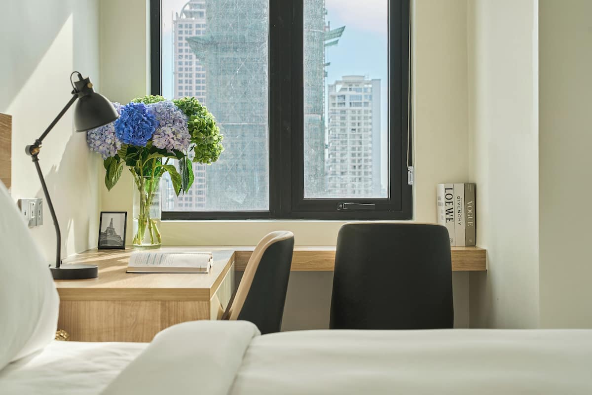 A hotel room with a bed, a desk with a lamp, a vase with flowers, and a view of tall buildings through a window.