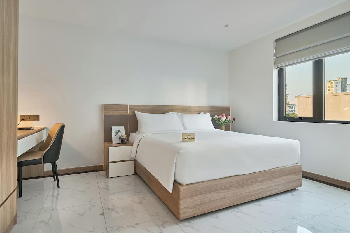 A modern hotel room with a large bed, a desk, and a chair. There is a window with a view of buildings outside.