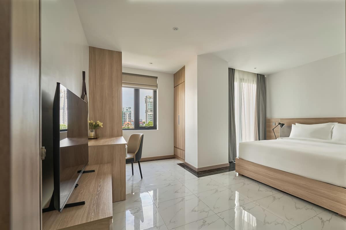 A modern hotel room with a large bed, a desk with a chair, a TV, and a window with a view of buildings.