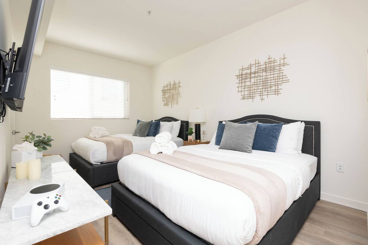 A modern bedroom with a large bed, a desk with a gaming console, and a TV mounted on the wall.