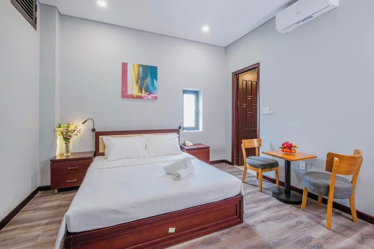 A bedroom with a wooden bed, a bedside table with a lamp, a small table with a chair, and a painting on the wall.
