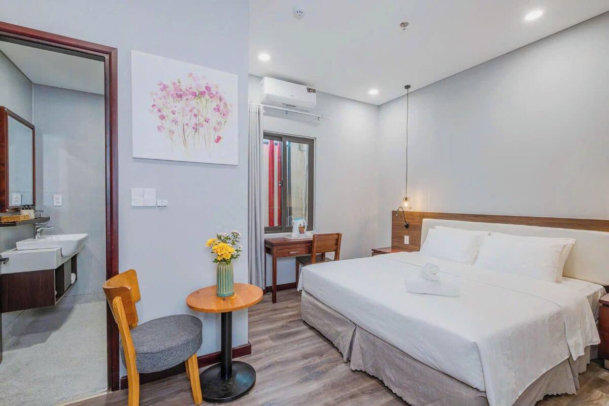 A hotel room with a bed, a desk, a chair, a small table with flowers, a mirror, and an air conditioning unit.
